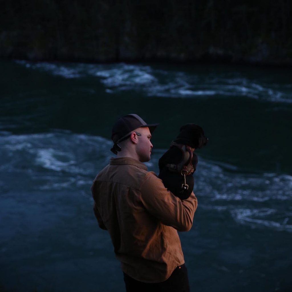 Country singer Zach Bryan stands in front of the ocean holding a dog