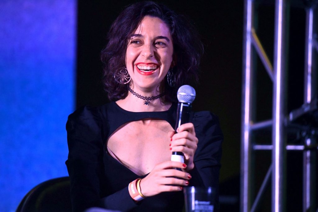 In an article about Jamie Loftus, the comedian stands against a blue and black background while holding a microphone.