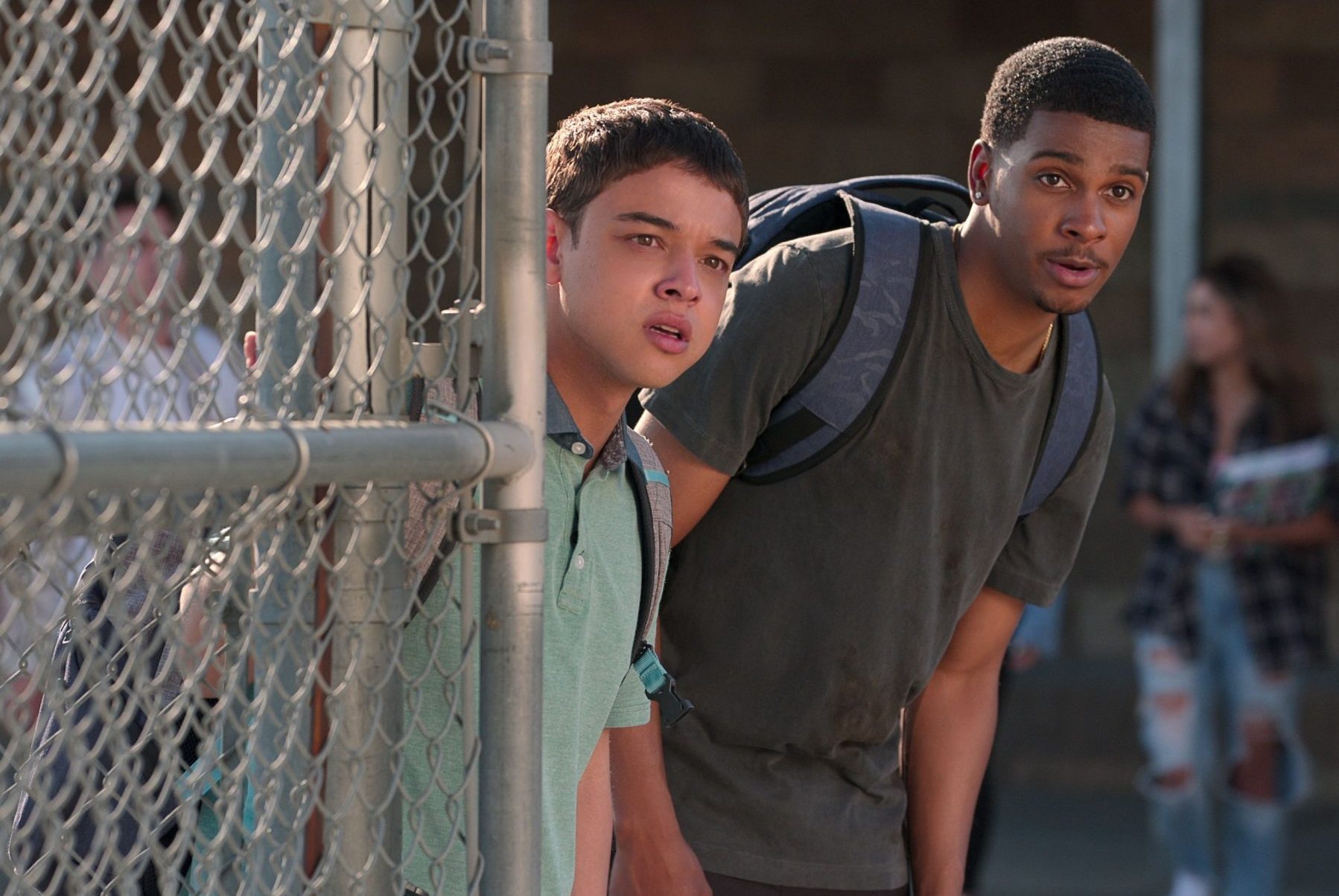 Two characters from Netflix's On My Block peek around a fence.