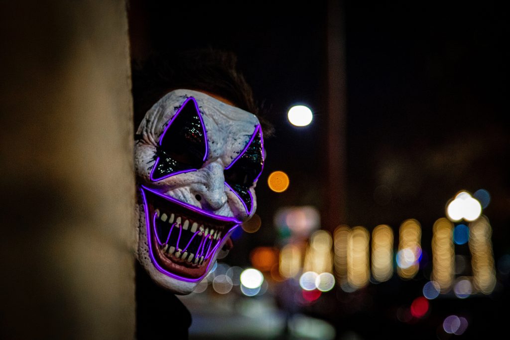 Horror character in scary clown mask peeking around the corner of a building at night