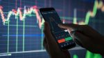 a phone with a trading app in front a computer with trading information