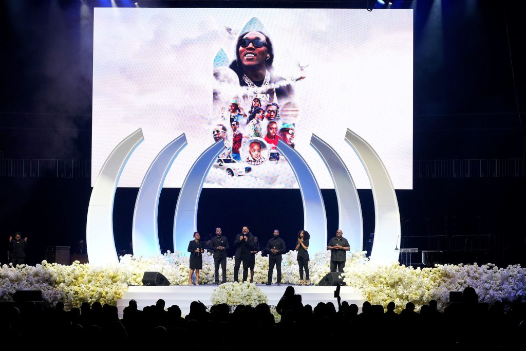 A screenshot from takeoff's funeral shows a group standing on stage in front of his picture on a screen.