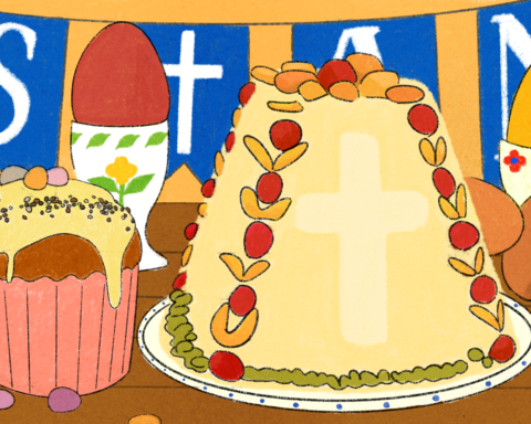 A series of desserts are set on a brown table before an orange background: a cupcake in a pink wrapper with yellow frosting and sprinkles, a yellow cake dotted with red and orange decorations and a cross drawn on it, candied eggs of red and yellow in white cups with painted markings. Bean shaped candies litter the table, while blue flags with white letters hang on top of the background.